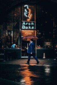 Man with Umbrella Walking by Restaurant Named Arch Duke