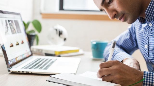 Man Writing in Notebook near a laptop