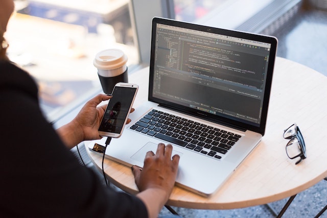 Person holding smartphone while working on laptop with coding