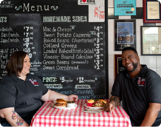 Two people sitting at a table with food in front of them and a chalkboard menu behind them