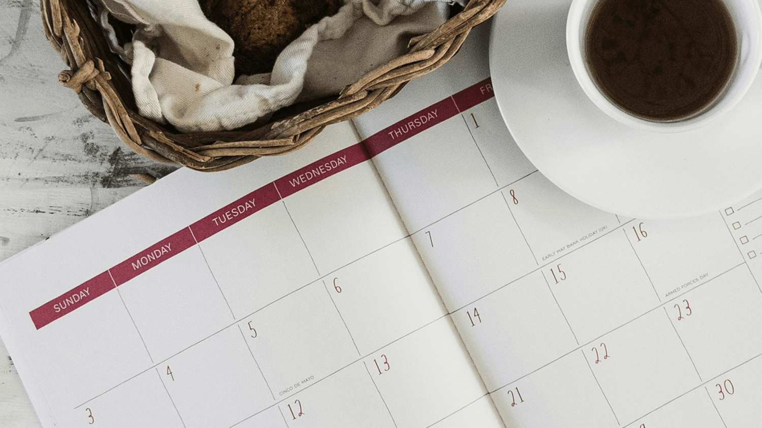 A calendar open to show the month spread with days of the week in red font. To the right, there is a cup of coffee on a saucer. The upper left corner of the image features a part of a wicker basket with a cloth and what appears to be a baked good inside. The background suggests a wooden surface.