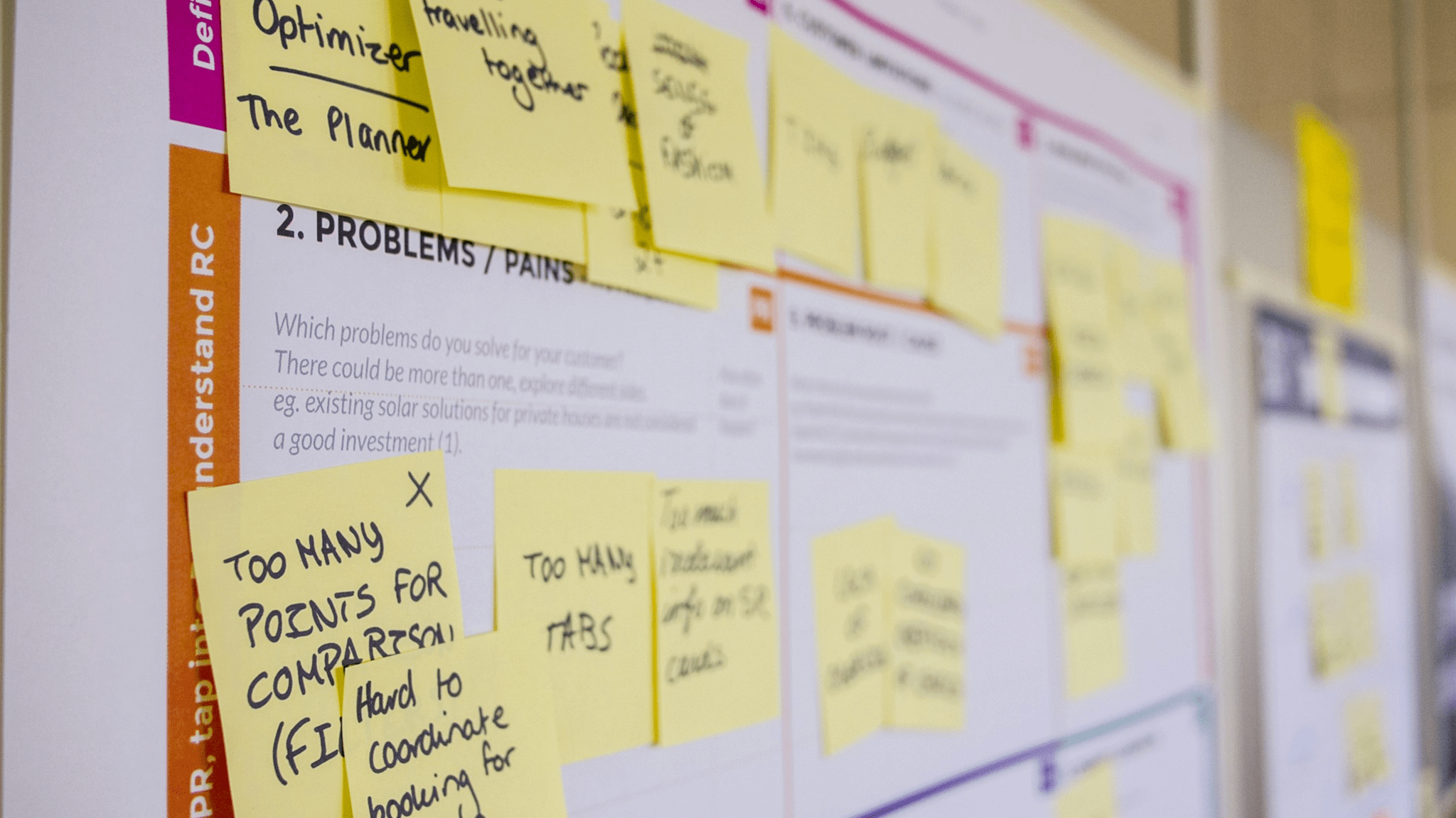 A close-up of a whiteboard covered with various yellow sticky notes containing handwritten text and a few sections highlighted with pink markers. The focus is on the foreground leaving the background slightly blurred, emphasizing the sticky notes