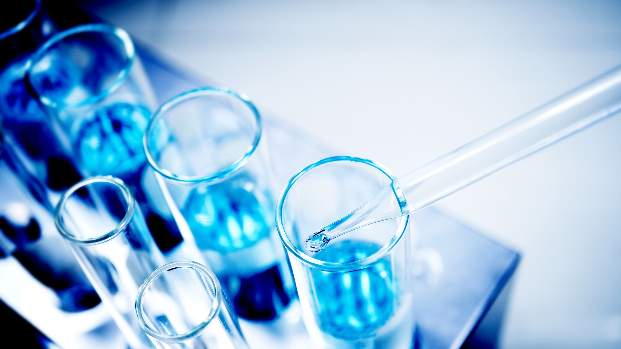 Close-up photo of a laboratory setting with a pipette dispensing a blue liquid into a test tube. Multiple test tubes filled with blue liquid are arranged in a holder, indicating a chemistry experiment or analysis in progress. The image has a cool blue tone, conveying a sterile, scientific atmosphere.