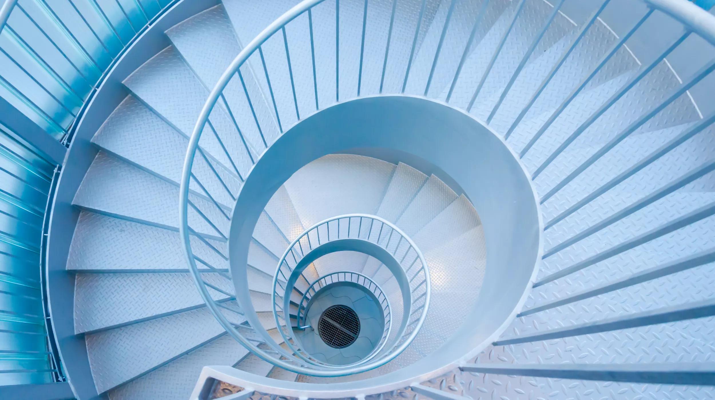 A top-down view of a spiral staircase with metallic blue hues, showcasing a symmetrical pattern as it descends floor by floor, culminating in a circular opening at the bottom.