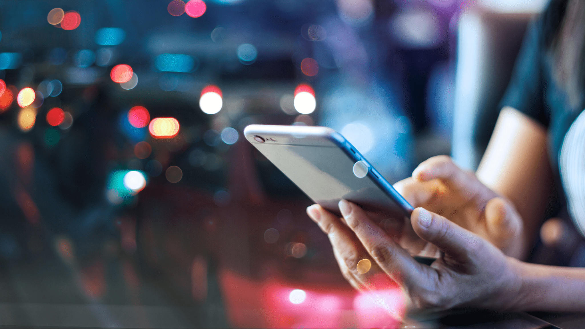 A person holding a smartphone with both hands against a blurred background of colorful city lights at night