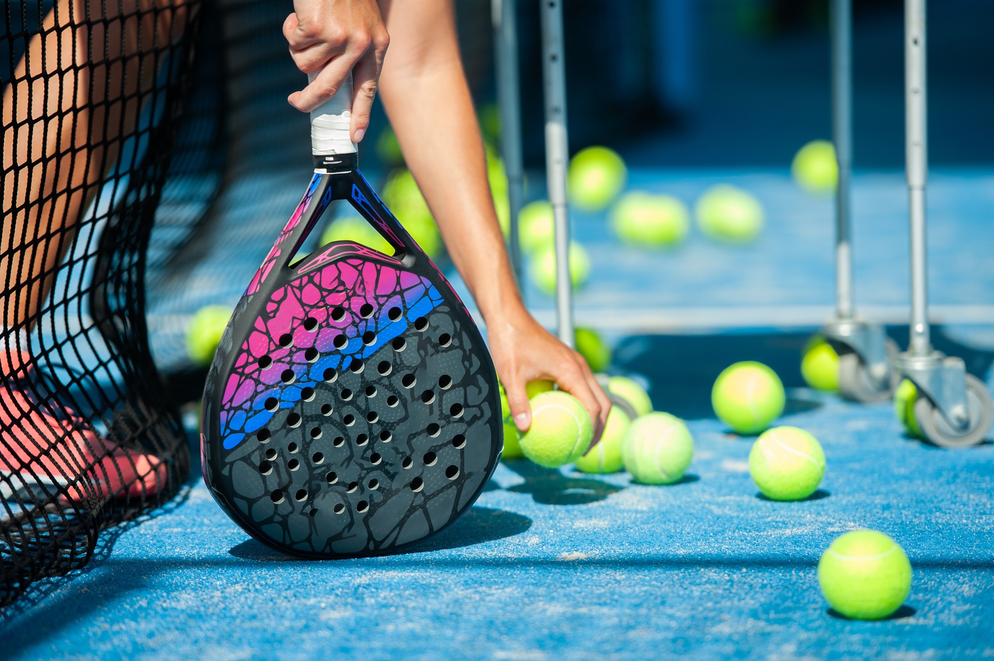 A close-up of a player's hand holding a padel racket with a pink and black design, picking up a yellow ball from a blue court surrounded by multiple other balls and a cart with wheels.