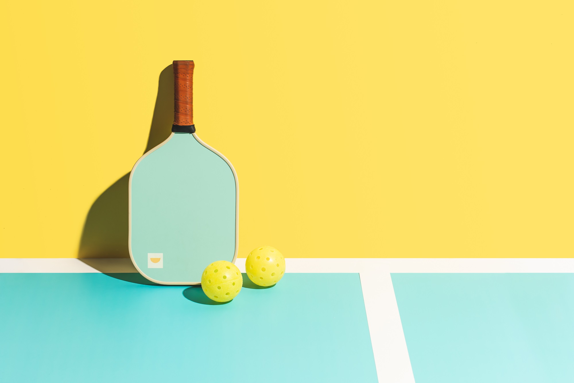 A pickleball paddle with a light blue playing surface and a brown handle stands upright against a bright yellow background, casting a shadow. Two yellow pickleball balls are positioned near the bottom of the paddle, resting on a pickleball court with blue area and white lines.