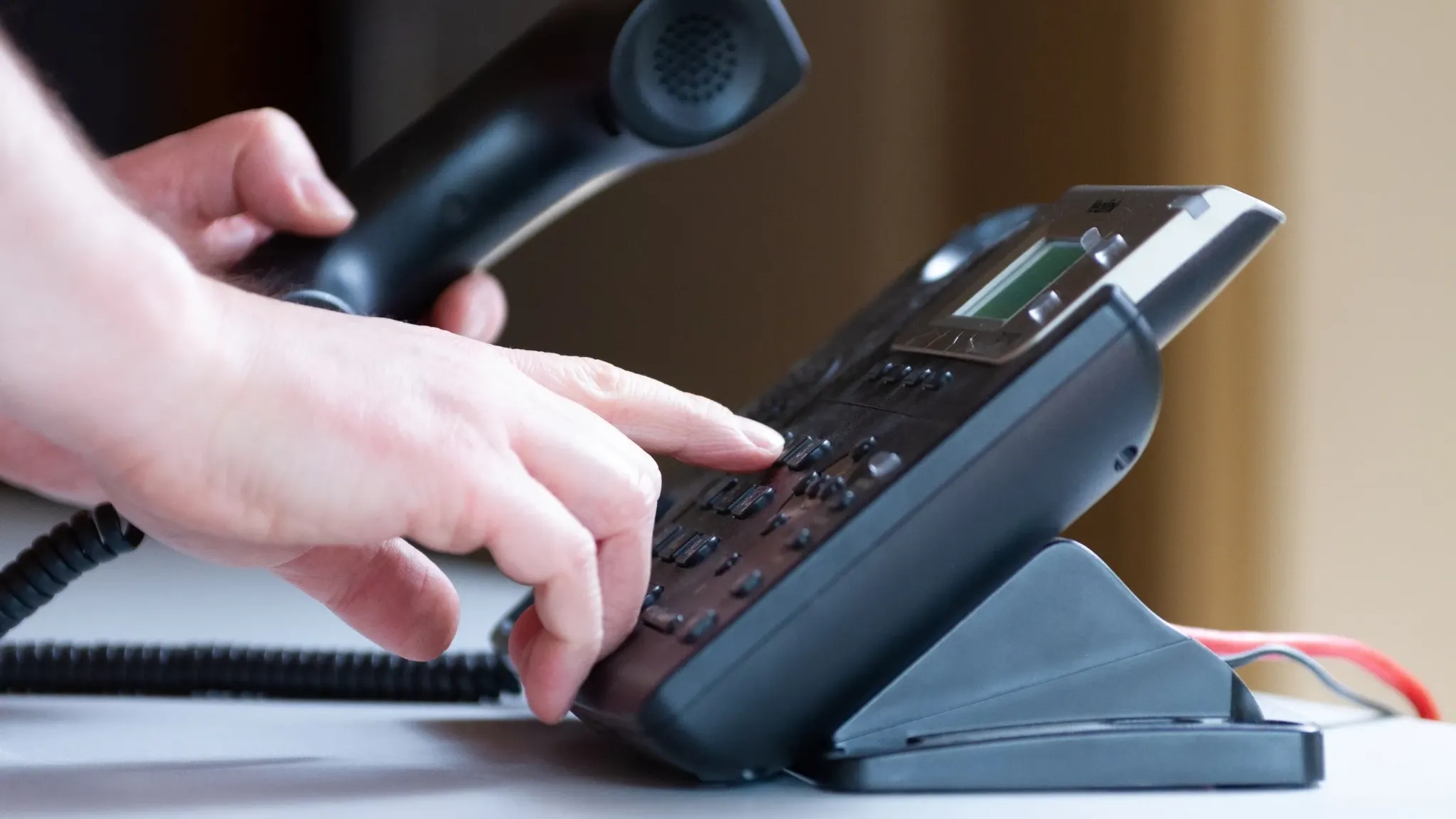 A person is holding a black phone handset in their left hand while dialing a number on a desk phone with their right hand. The phone is positioned on a stand, and the image focuses on the hands and the phone, capturing the act of making a call.