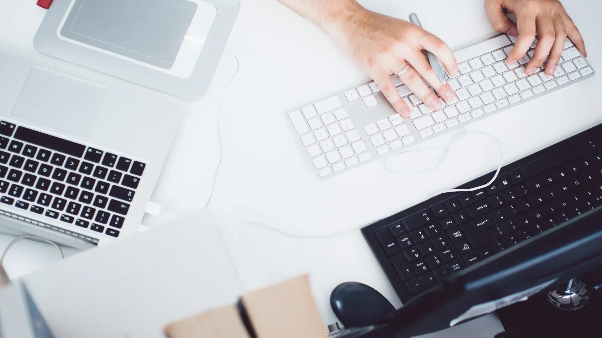 A top-down view of a workspace featuring a person's hands typing on a white keyboard. Nearby, there is a laptop with a black keyboard, a graphics tablet with a stylus, and a black keyboard. The scene is set on a white desk, creating a clean and organized environment.