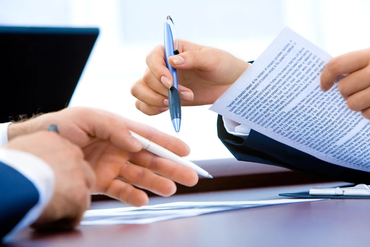 Two Hands Hold Pens and Printed Paper above a Desk