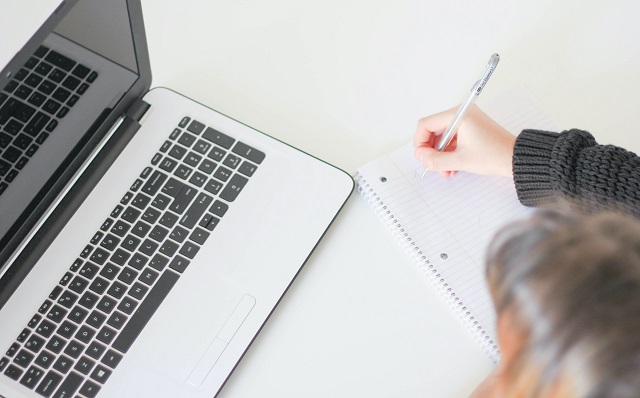 Woman Writing In Notepad Near Laptop On Desk