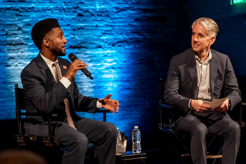 Mayor Scott holding a microphone, seated next to and speaking with Jeremy Hartman, on stage in at the GoDaddy Open Baltimore event